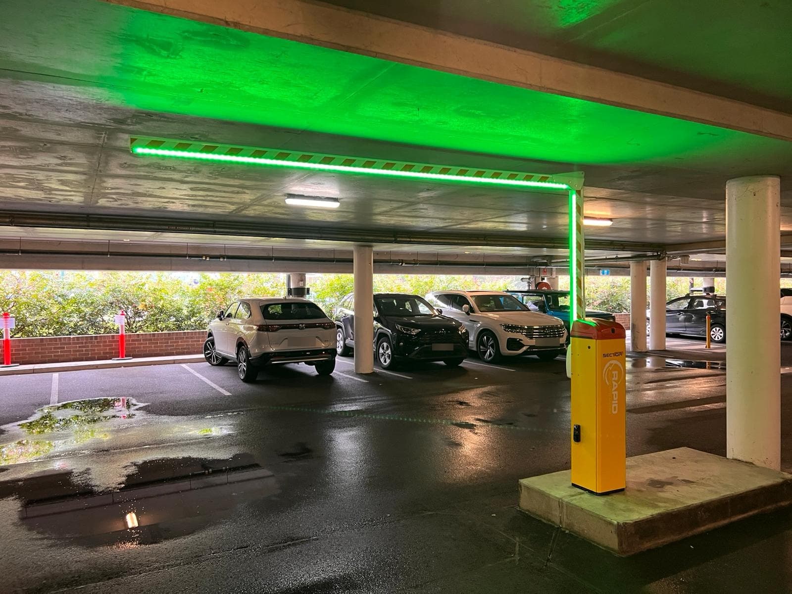 Operational boom gate beneath green lane guidance lighting in a covered car park.