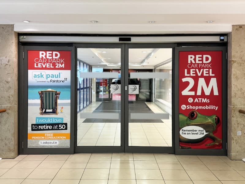 Automatic sliding glass doors at a shopping centre entrance featuring advertisements and directional information on either side.