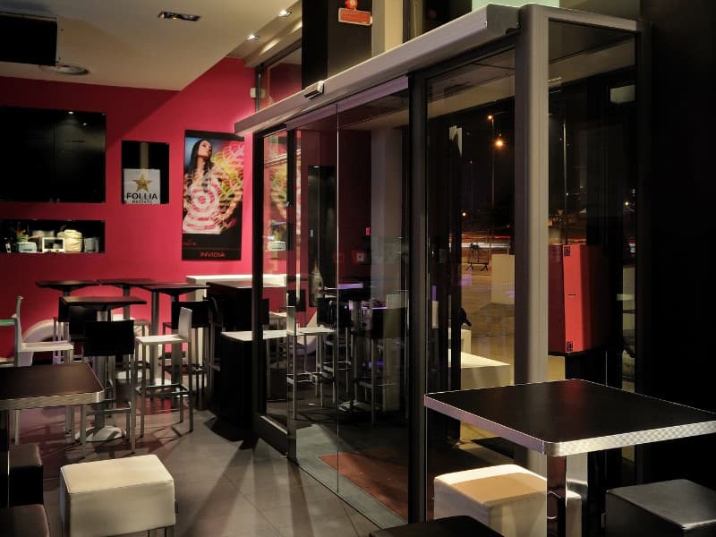 Automatic sliding glass doors at the entrance of a modern café with a stylish interior, featuring black and white furniture, vibrant wall art, and ambient lighting.