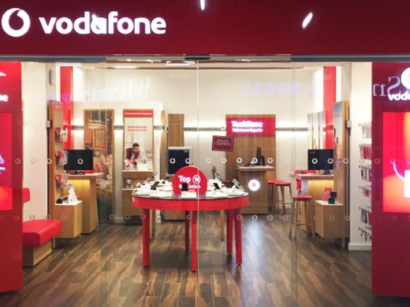 Front view of a Vodafone retail store with automatic sliding glass doors, showcasing a modern interior with phone displays, red branding, and wood flooring.