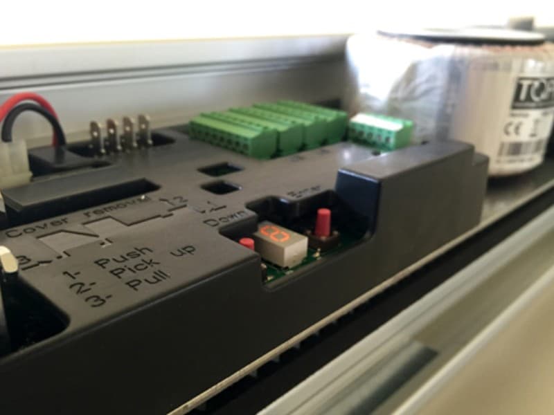 Detailed close-up of an automatic door control panel, featuring connection terminals, green input modules, a digital display, and operational buttons for configuration.
