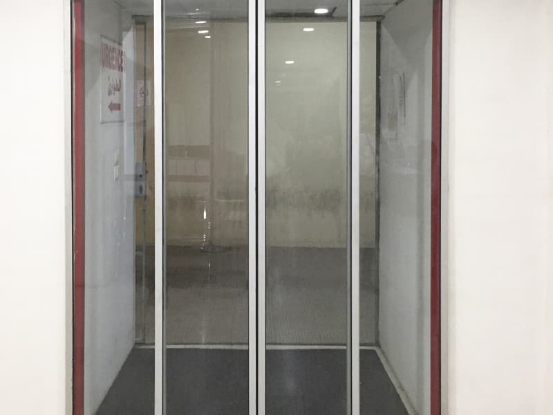 Frontal view of a three-panel automatic telescopic sliding glass door in a commercial hallway, framed by white walls and overhead recessed lighting.