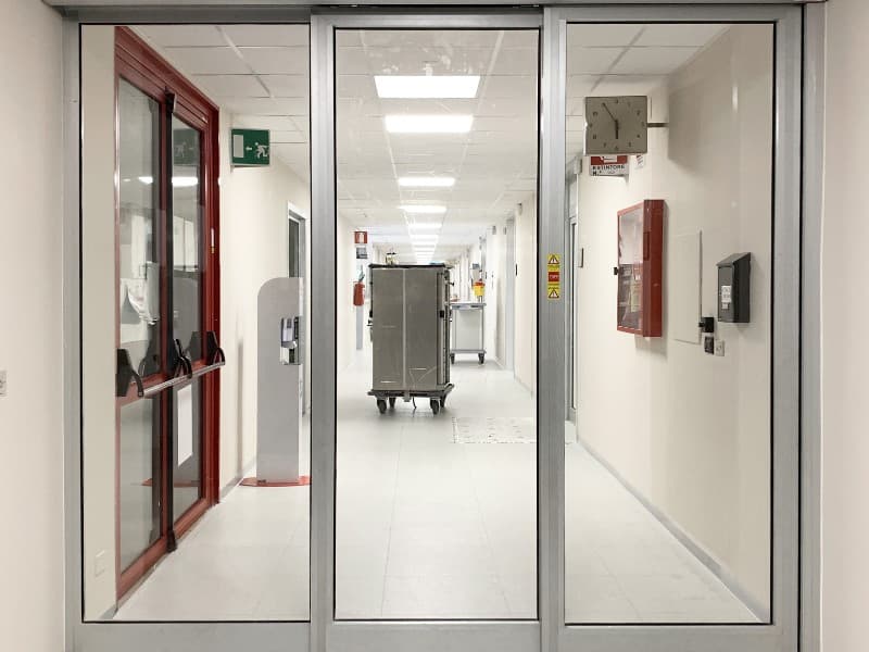 Automatic sliding glass doors in a sterile hospital corridor, revealing medical carts and equipment in a clean, well-lit hallway with white walls and tiled flooring.