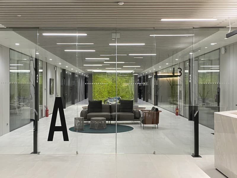 Modern office lobby with large frameless glass sliding doors, featuring minimalist decor, lounge seating, and a green wall backdrop at the end of a well-lit corridor.