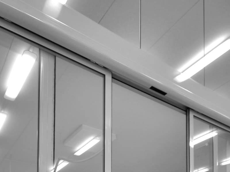 Automatic sliding glass doors in a clean, modern facility with overhead motion sensor and white ceiling panels illuminated by fluorescent lighting.