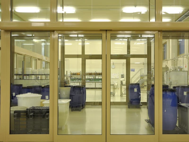 Automatic sliding glass doors with an aluminium frame providing access to a clean industrial facility, with large blue barrels and stainless steel equipment visible inside.
