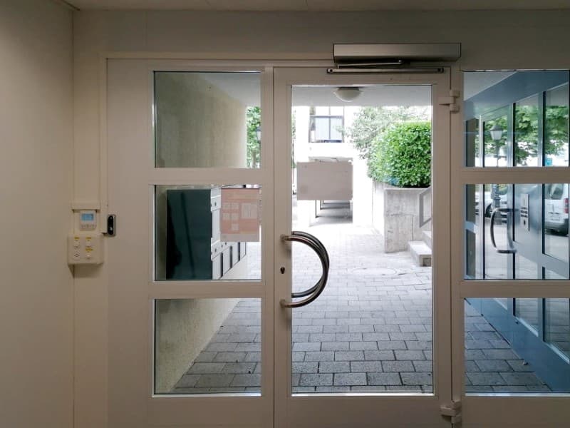 Glass double entrance door with circular handles, integrated with an overhead automatic door closer and access control panel mounted on the left wall, leading to an outdoor pathway.