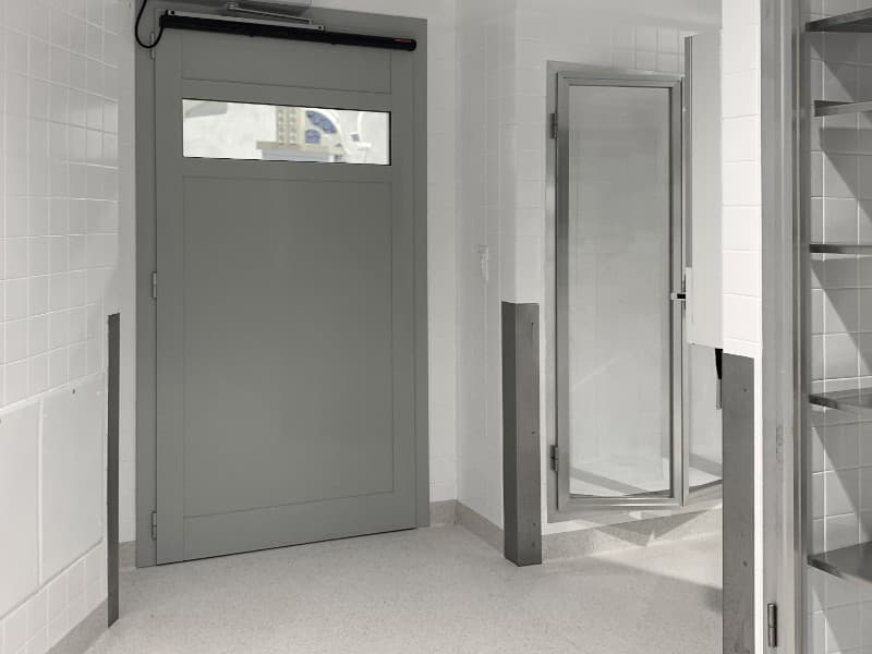 Industrial-style grey swing door fitted with an automatic door operator in a sterile, tiled environment, likely within a medical or laboratory facility.