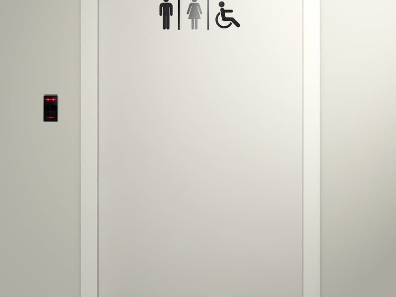 Minimalist automatic sliding door with gender-neutral and accessible restroom signs above, and a black card reader with red indicator lights mounted on the wall to the left.