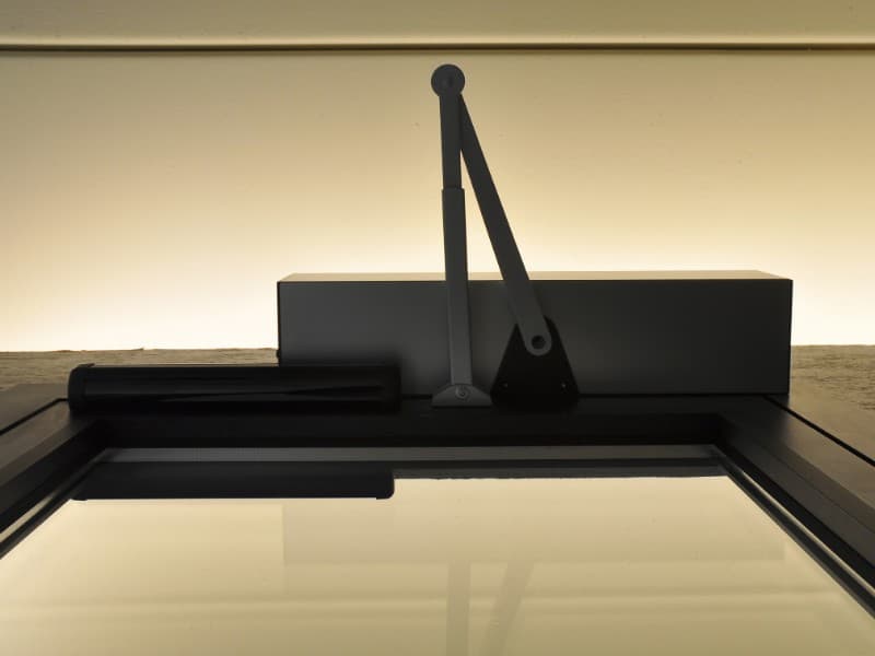 Close-up view of an automatic door closer mechanism installed on top of a glass door, with ambient lighting in the background.
