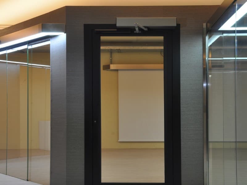 Modern glass door with an automatic swing door operator installed at the top, situated in a sleek commercial or office interior with glass partitions and illuminated ceilings.