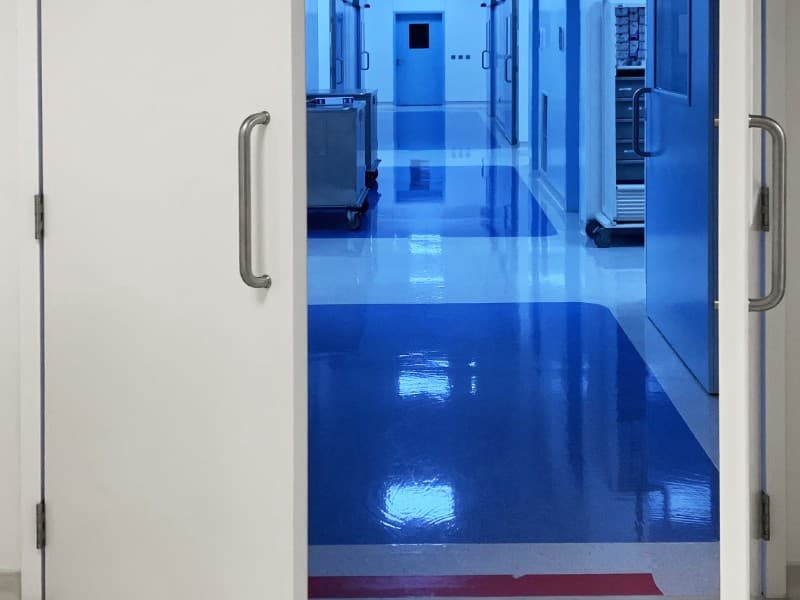 Cleanroom hallway with highly polished blue and white flooring, viewed through open double swing doors with stainless steel handles, in a sterile industrial or laboratory environment.