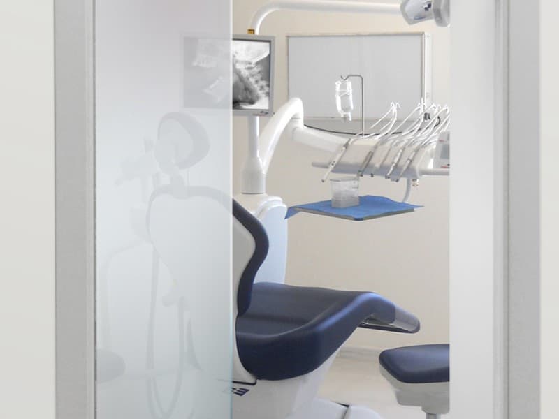 View through a frosted glass sliding door into a clean, modern dental clinic room with a blue dental chair, equipment tray, and mounted monitor displaying an X-ray.