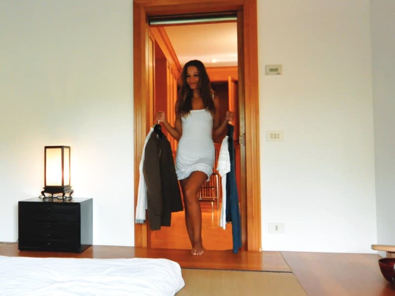 Person entering a modern bedroom through a wooden-framed automatic sliding door, holding clothes in both hands, with warm ambient lighting in the background.