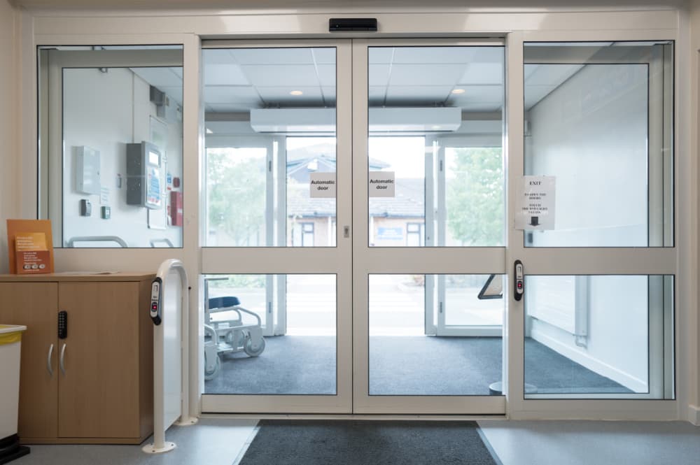 Interior view of a modern hospital entrance with automatic sliding glass doors, showcasing clear signage, access control panel, and a wheelchair near the exit.