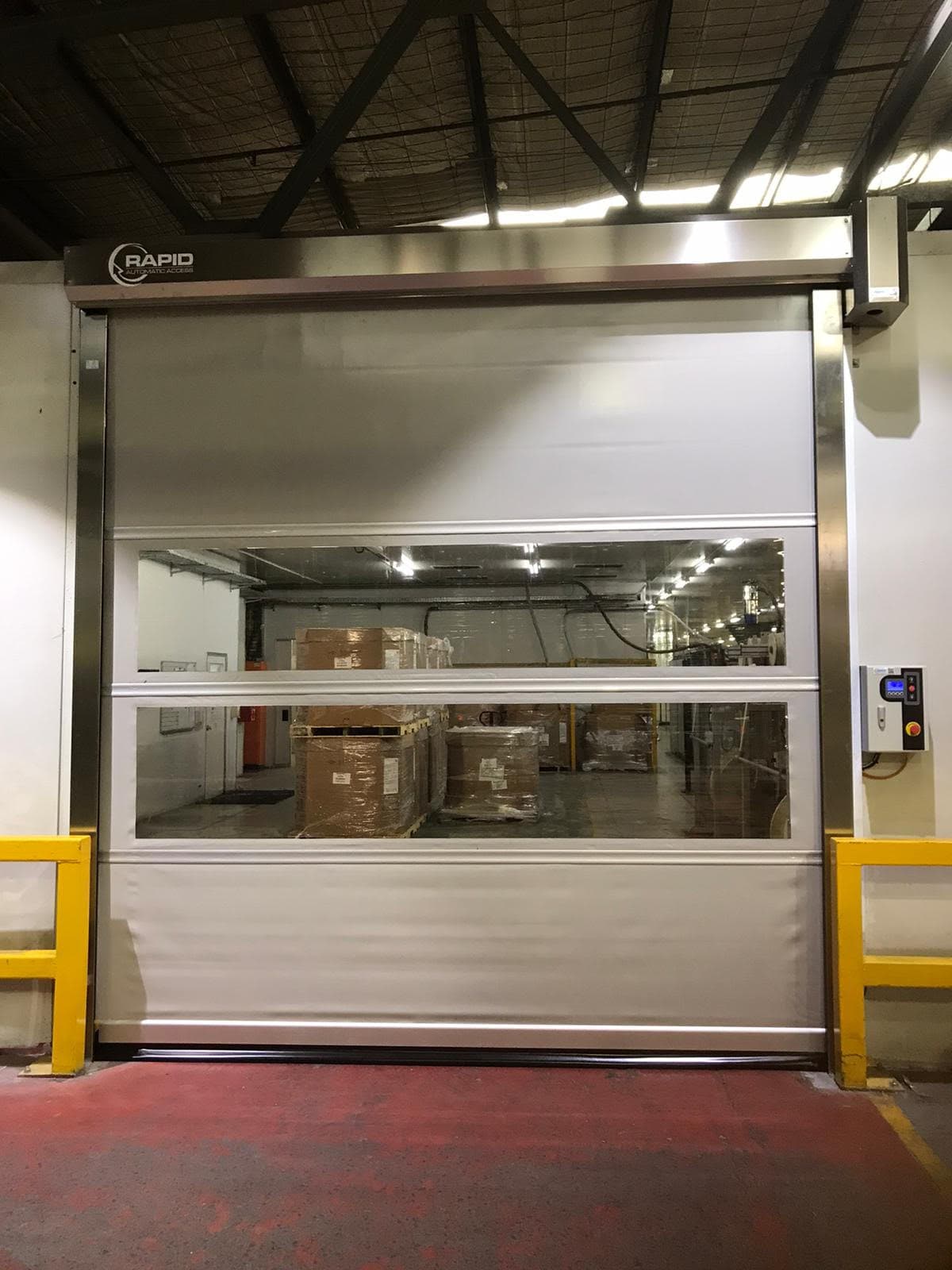 White high-speed door with two transparent vision panels showing stacked cardboard boxes in a warehouse interior. The door frame has yellow safety barriers on both sides, and the Rapid logo is visible on the top header. A red floor marking is visible at the base.