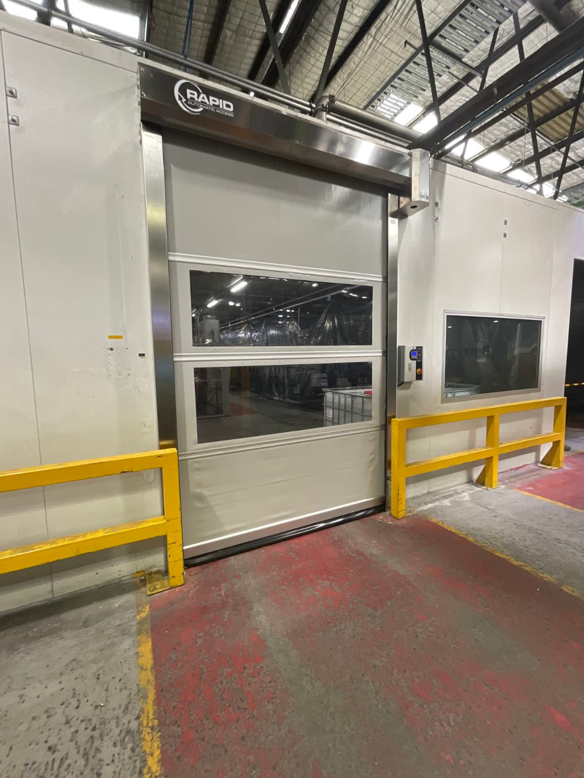 Red high-speed roller door with illuminated white LED safety strips across the middle. The door has a stainless steel frame, with control panels mounted on both sides and yellow safety railings visible to the left.