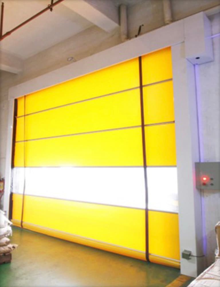 Photo of a yellow high-speed roller door with a white transparent vision panel in the middle, installed in an industrial space with green epoxy flooring. A red control panel is mounted on the right side of the doorway.