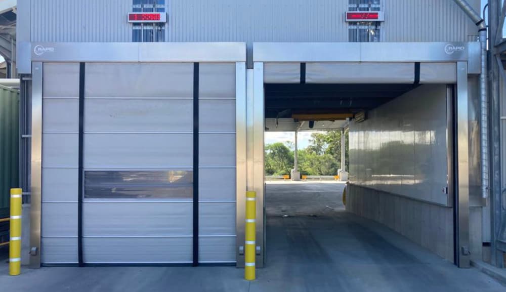 Photo of two white high-speed doors installed side by side in an industrial loading dock area, one closed and one open, with yellow safety bollards and red digital displays mounted above. The open door shows a concrete ramp leading to the exterior.