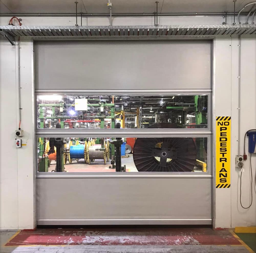 Photo of a high-speed door at cables depot with transparent vision panels showing industrial cable spools and manufacturing equipment. A yellow 'NO PEDESTRIANS' safety sign is mounted on the right side, and the doorway has red and yellow floor markings.