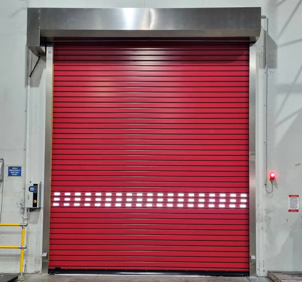 Photo of a red high-speed roller door with illuminated white LED safety strips across the middle. The door has a stainless steel frame, with control panels mounted on both sides and yellow safety railings visible to the left.