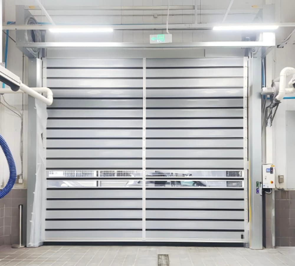 Photo of a white high-speed door with horizontal slats and a transparent vision panel installed in a clean room environment. The door features stainless steel side columns, overhead fluorescent lighting, and ventilation ducts visible on both sides.